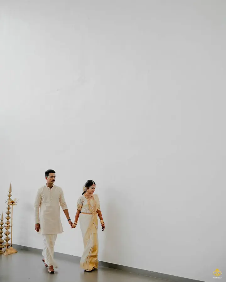 Kerala couple in cream and gold kasavu walk hand in hand past a lit nilavilakku
