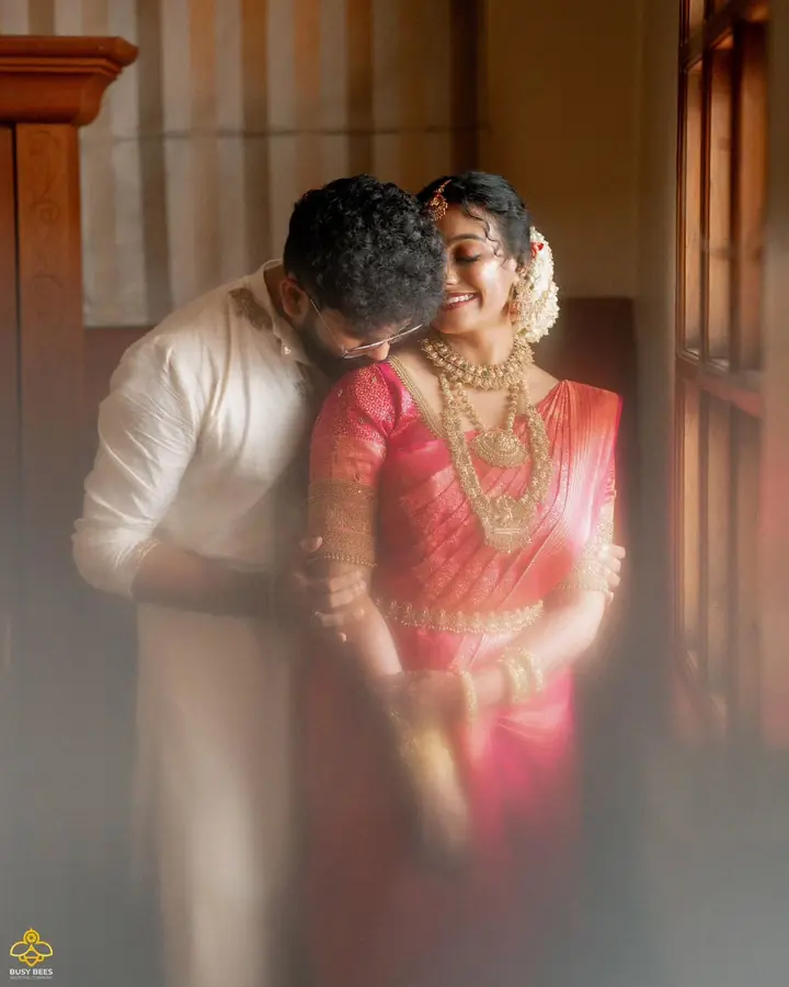 Bride and groom hold each other in soft window light, in pink and cream kasavu