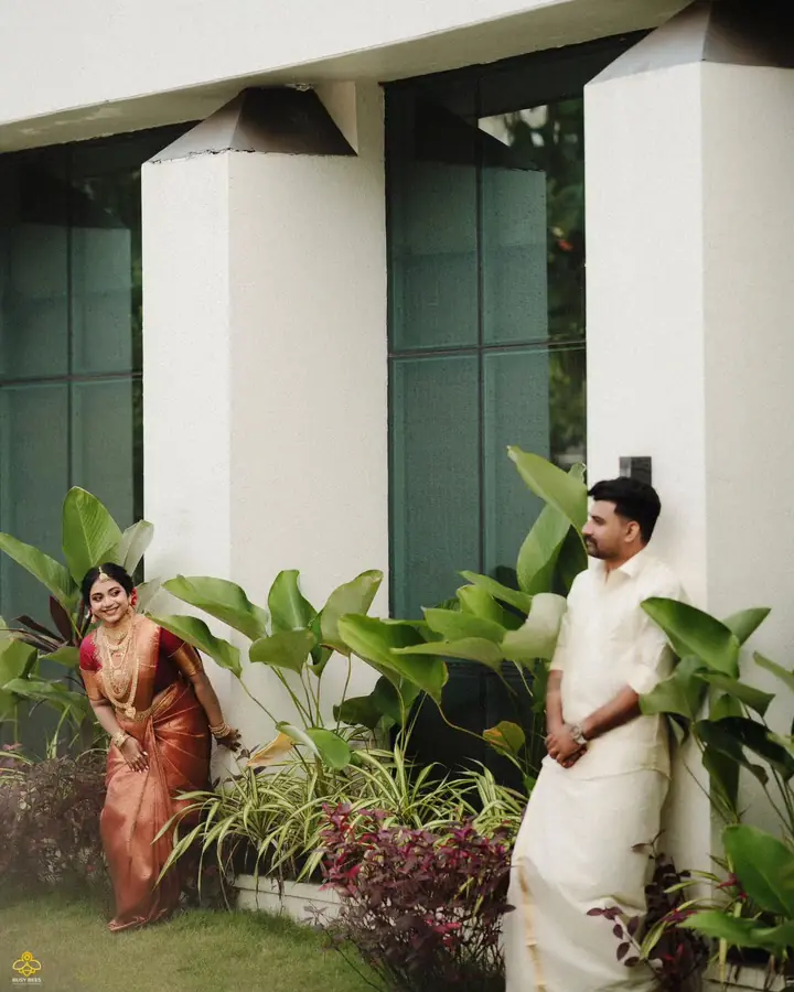 Bride in a maroon silk saree and groom in a white mundu in a garden courtyard